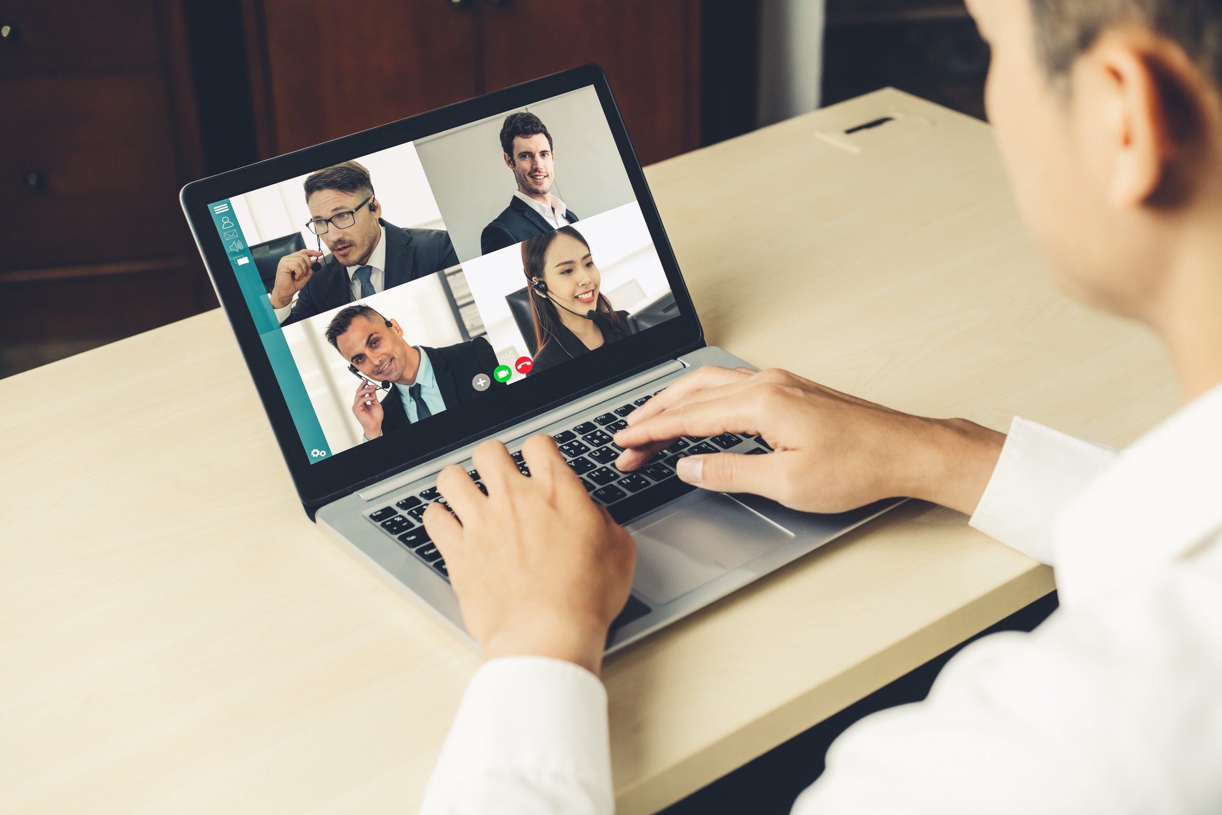 A person participating in a video conference on a laptop busy with a remote deposition, with five individuals displayed on the screen, each wearing business attire. The participant is typing on the keyboard, seated at a wooden desk in a professional setting.