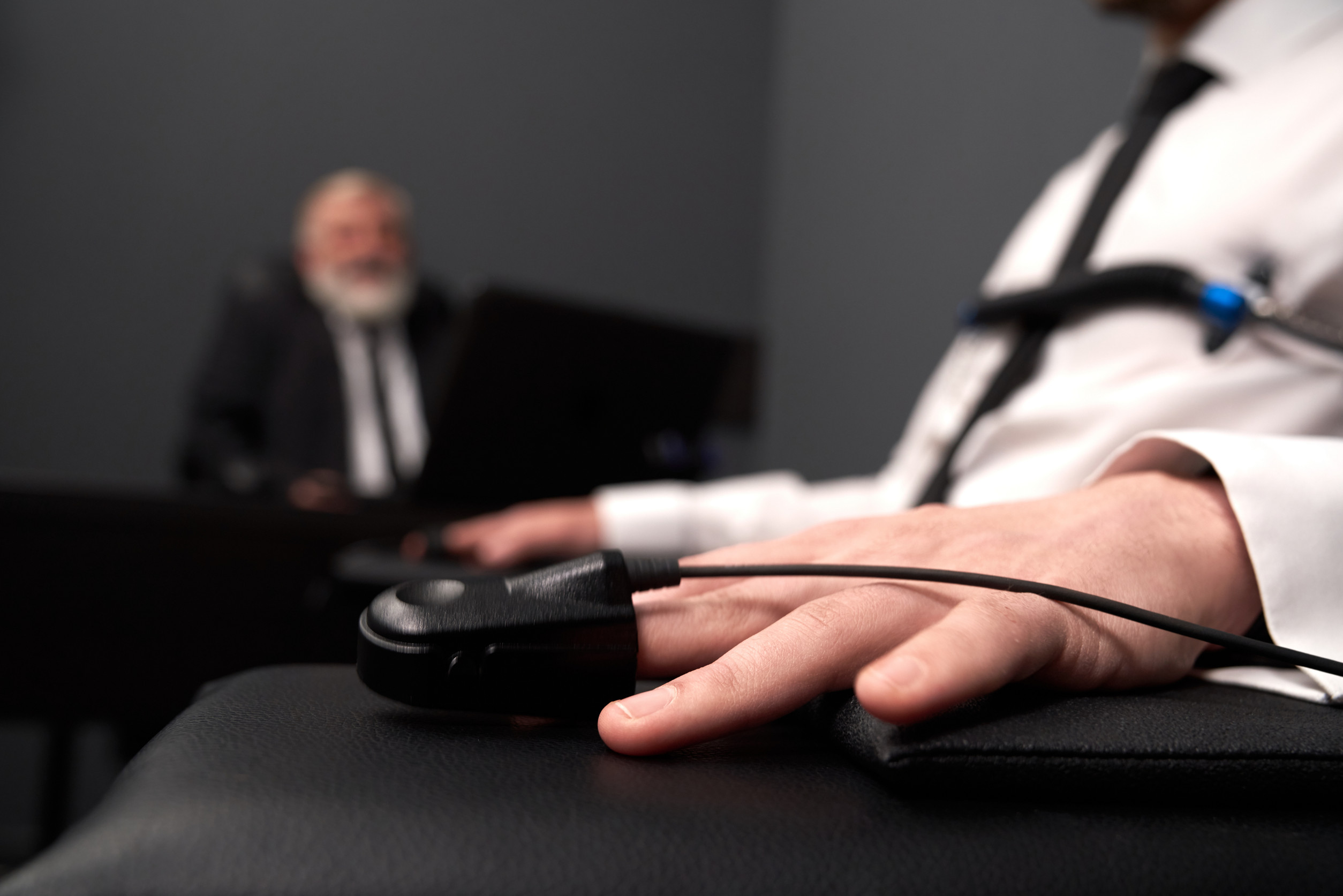 A person undergoing a polygraph test with sensors attached to their hand and chest, sitting in a formal environment. In the background, a blurred examiner in a suit observes the process, creating a serious and investigative atmosphere.