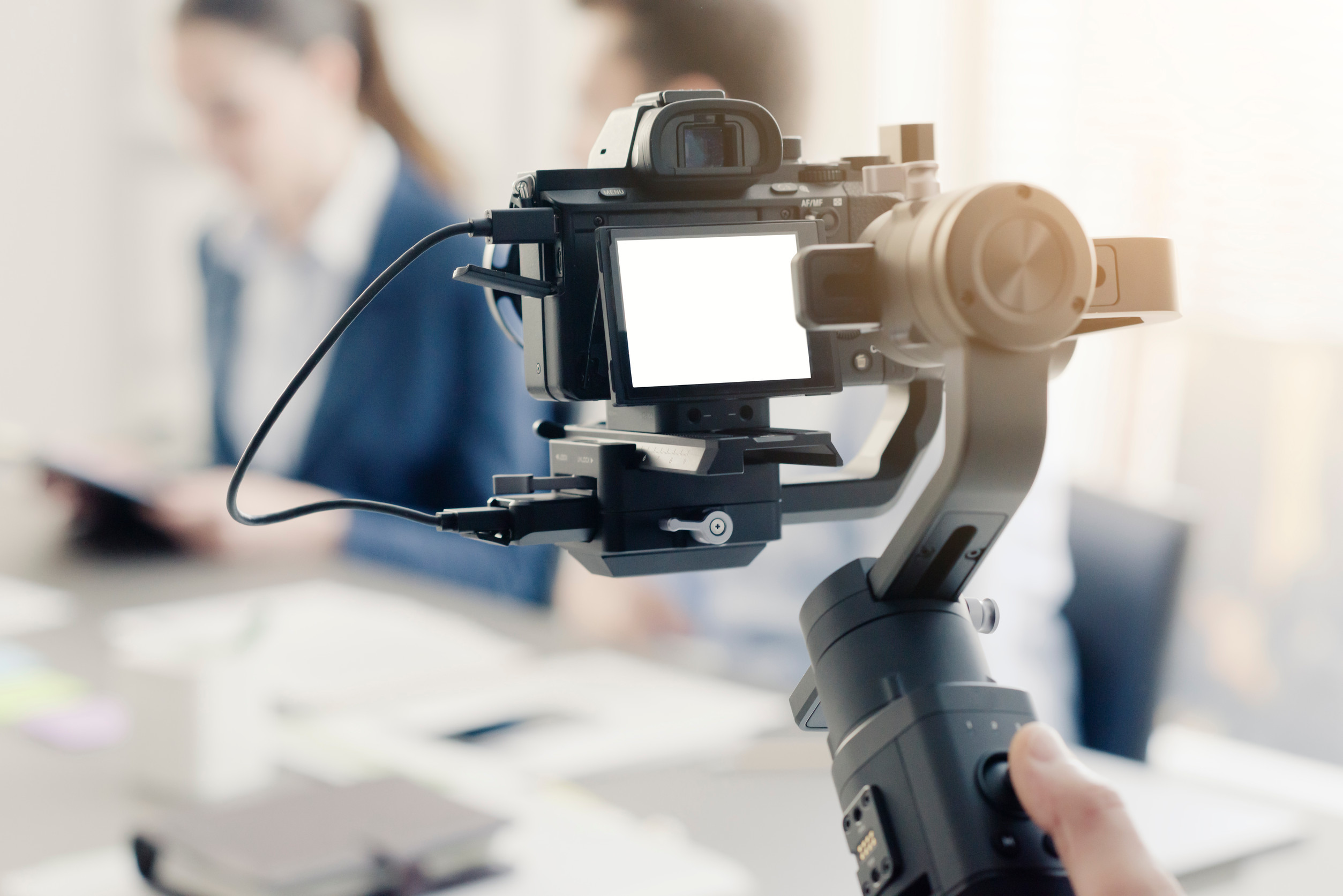 A close-up view of a professional video camera mounted on a stabilizer gimbal, capturing footage in an office setting. In the background, two blurred individuals are engaged in a business discussion, creating a professional and focused environment.