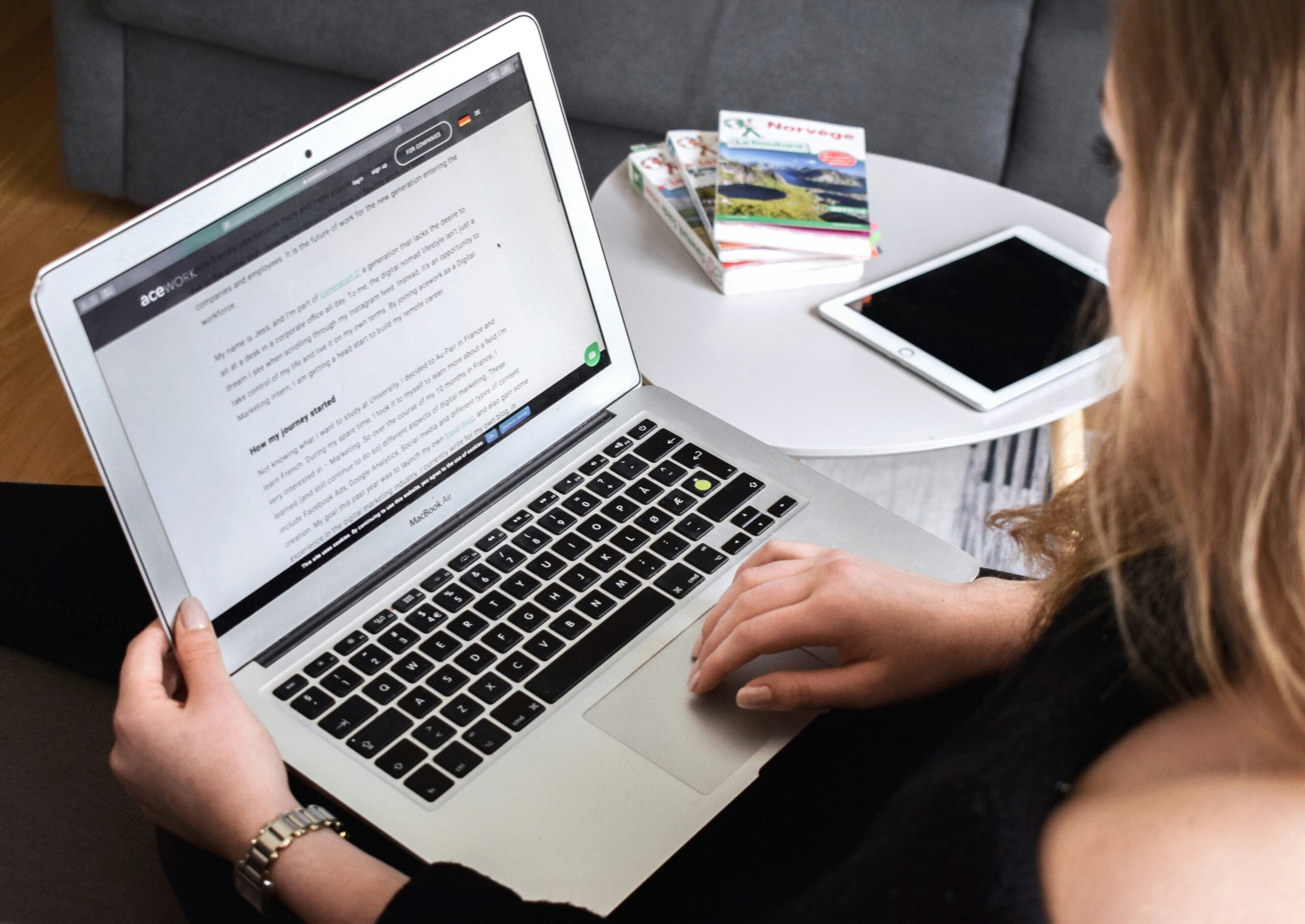 A woman using a MacBook Air laptop, reading an article on the screen. She is sitting on a couch, with a white round table nearby holding an iPad and a few travel guidebooks. The setting suggests a relaxed learning or remote work environment using AI in legal practice terms.