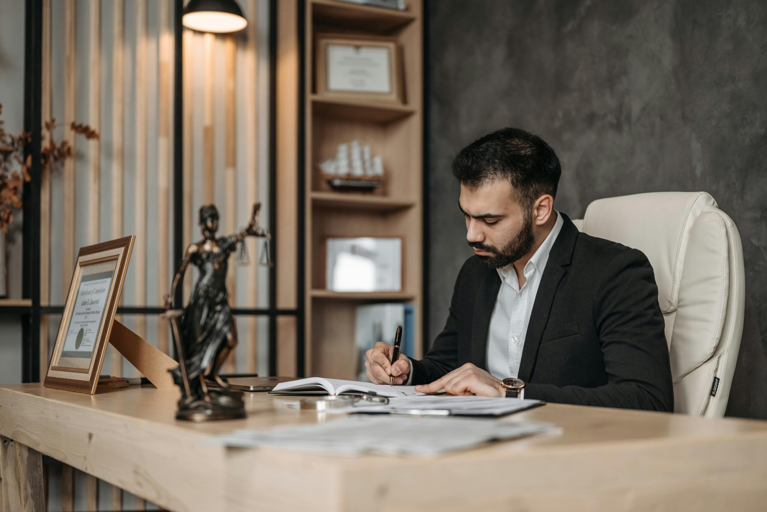 A focused lawyer in a black suit sits at a wooden desk, writing on documents in a modern office. The desk features a bronze Lady Justice statue, a framed certificate, and stacks of paperwork. Behind him, a bookshelf with legal books and decor adds to the professional setting. Remote deposition on legal practice.