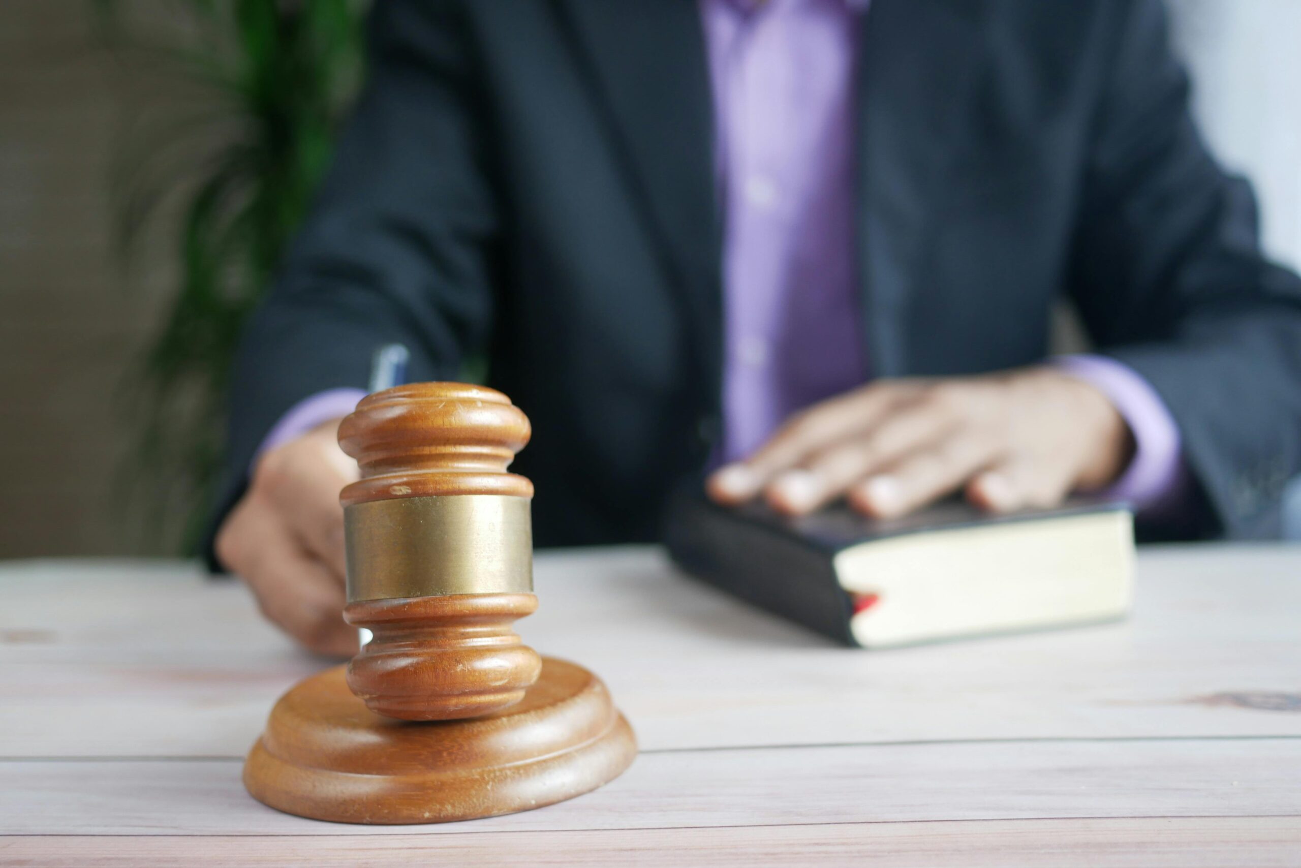 A close-up of a wooden gavel on a desk, with a judge or lawyer in a dark suit and purple shirt in the background. One hand is holding a pen, while the other rests on a thick legal book, symbolizing justice, law, and courtroom decision-making.